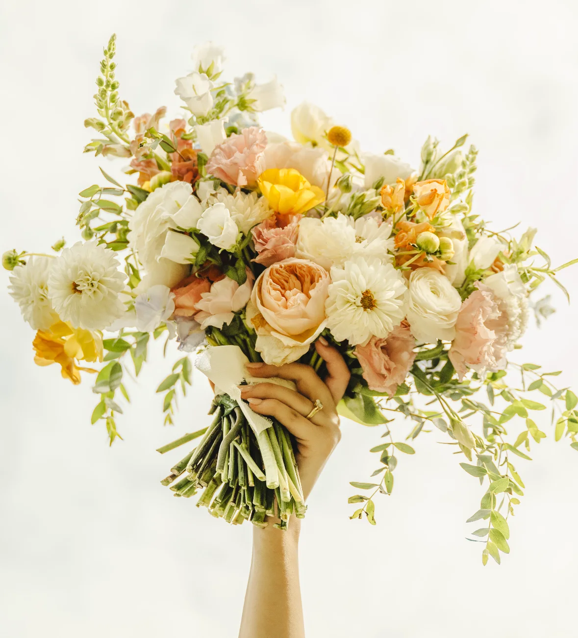 A bride holding up a bouquet of cream, peach and yellow blooms with trailing greenery